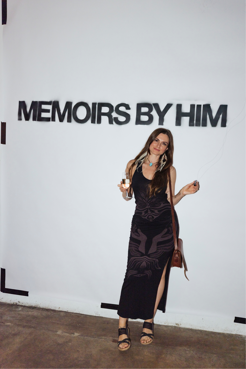 Woman in a black dress standing in front of a wall with 'Memoirs by Him' text.