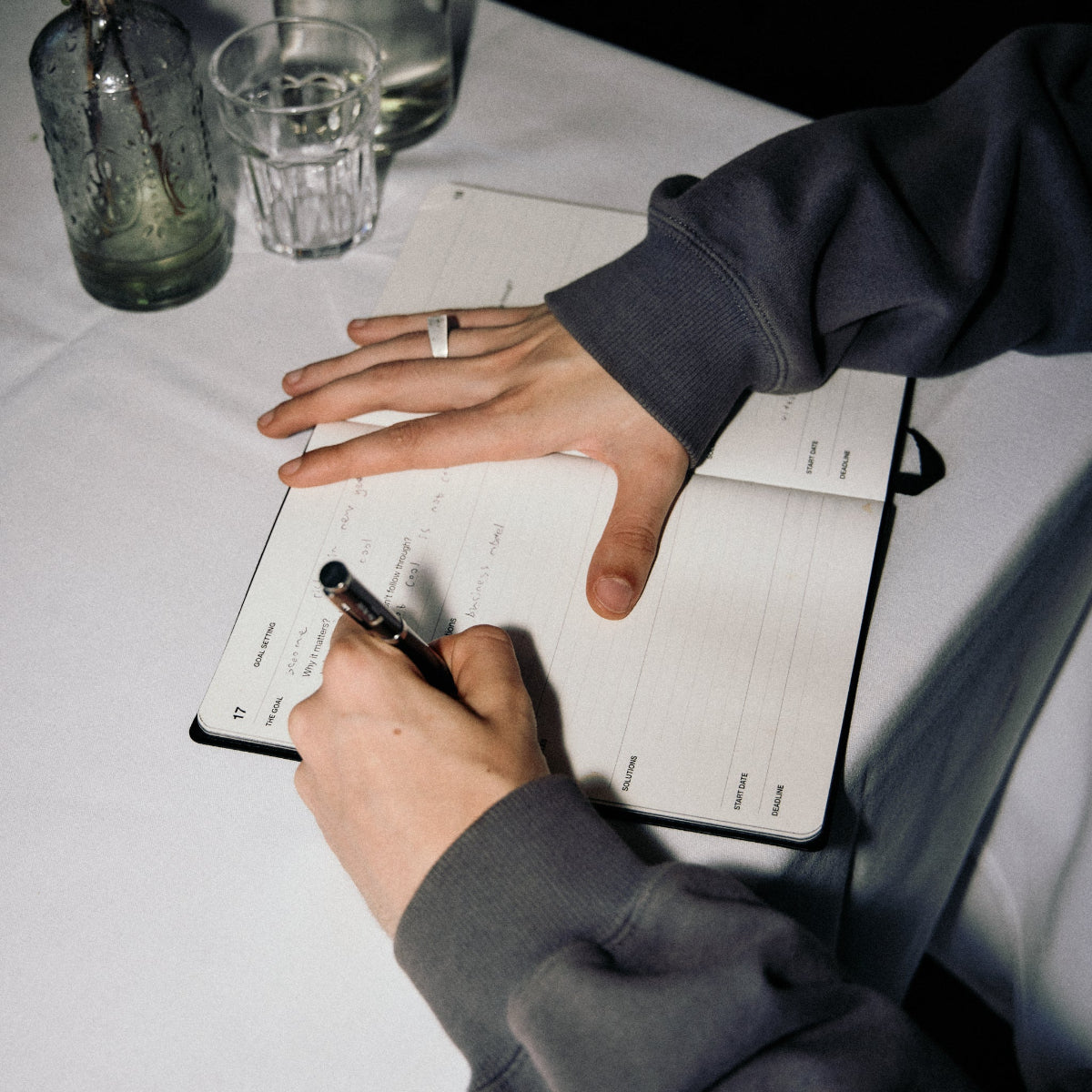 Person writing in a notebook with a pen on a white tablecloth.