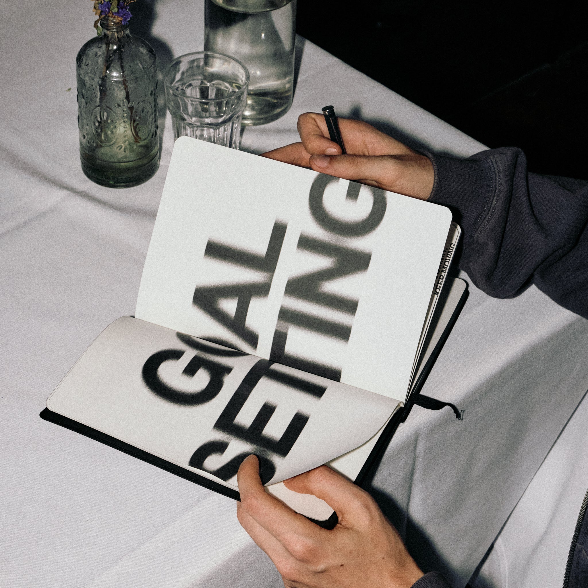 Person holding a journal with 'GOAL SETTING' text on a table with glasses and a bottle.